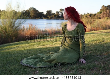 full length portrait of a red haired beautiful lady wearing a green medieval gown near a lake. Royalty-Free Stock Photo #483951013