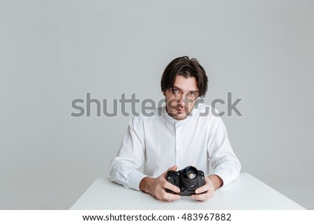 Handsome young brunette man sitting at the table holding camera isolated on the gray background