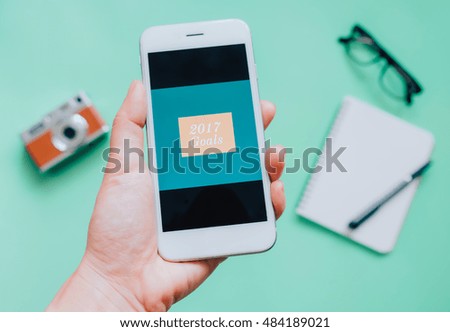Hand holding smartphone and 2017 goals on screen with camera, notebook and eyeglasses on green background