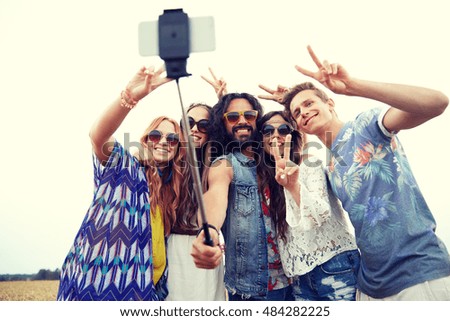 nature, summer, youth culture, technology and people concept - smiling young hippie friends in sunglasses taking picture by smartphone on selfie stick and showing peace gesture on cereal field