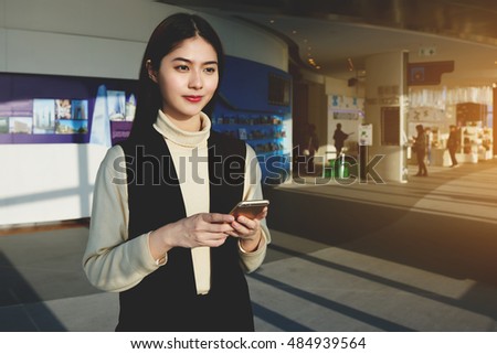 Chinese woman CEO is waiting for an answer on her text message on mobile phone, while is standing in airport terminal before her business trip abroad. Hipster girl is using cell telephone for chat