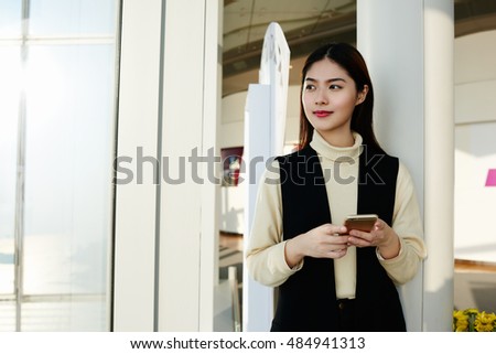 Young Asian woman successful economist is waiting for a call from her college, while is standing in hallway office building near copy space for your advertising text message or promotional content