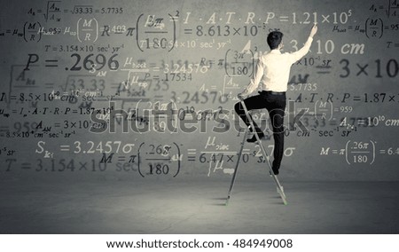 A businessman in elegant suit standing on a small ladder and writing numbers, calculating on grey wall background