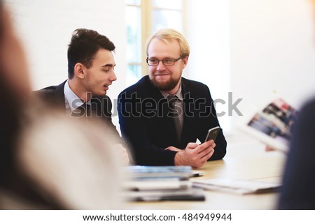 Man professional manager is showing mobile phone his partner, while they are sitting in modern office interior. Two men are watching news in internet via cell telephone, while are waiting interview