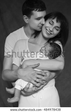 Little infant baby boy sleeping laying on mothers and fathers arms. Mother and father smiling. Neutral black background, black and white picture. All in white clothes. Happy family