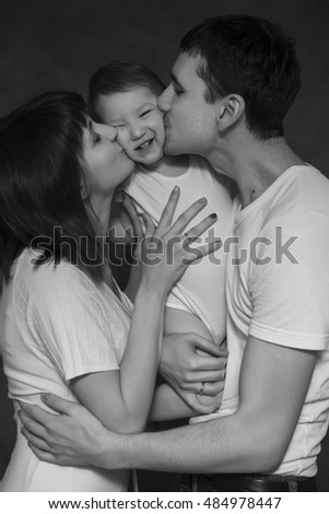 Little cute 2 years girl laying on mothers and fathers arms smiling, they are kissing her. Neutral black background, black and white picture. All in white clothes. Happy family