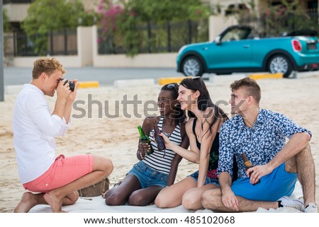 Friends and beer. Four young cheerful people taking photo and  cheering with beer and smiling while sitting on the blanket on a beach in front of a car and  villa