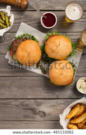 Burgers and cheeseburger on wooden table with sauces, fries, pickles and lager beer, overhead view with copy space