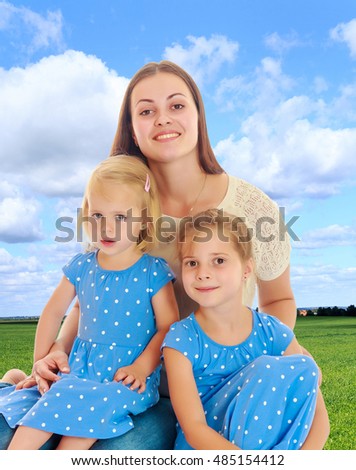 Beautiful young mother with her two daughters. Girls in the same dress with polka dots. Look directly at the camera.On the background of green grass and blue sky with clouds. 