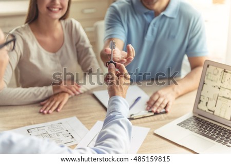 Cropped image of handsome realtor in classic shirt giving key of the new apartment to happy young couple while they are sitting in the office