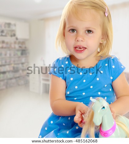 Cute little blonde girl playing with a toy horse. Girl wearing a blue dress with polka dots.On the background of the room where children live, and are on the shelves of toys.