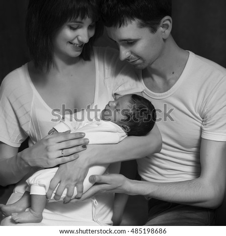 Little infant baby boy sleeping laying on mothers arms. Mother kissing father. Neutral black background, black and white picture. All in white clothes. Happy family
