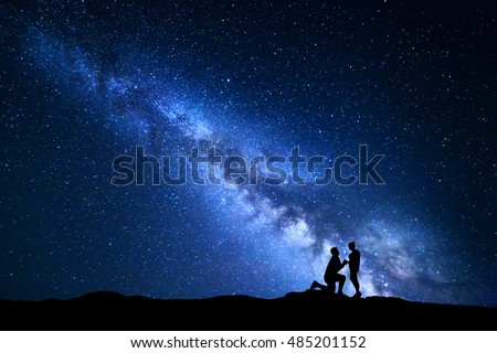 Milky Way. Night landscape with silhouettes of a man making marriage proposal to his girlfriend and starry sky. Silhouette of lovers. Couple, relationship. Blue Milky way with people. Universe