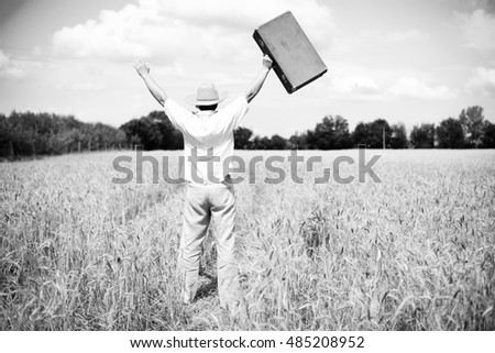 Black and white back view picture of happy man holding suitcase rising hands in wheat field. Back view of male excited on countryside background.