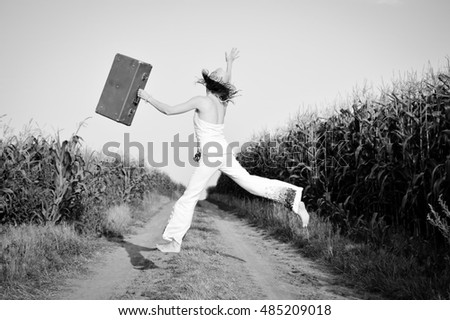 Black and white picture of jumping girl wearing straw hat with suitcase in corn field. Back view of happy joyful woman on the road outdoors background