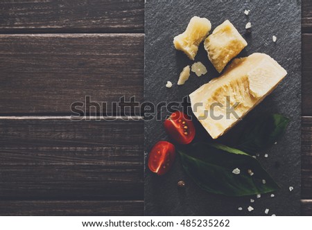 Cheese delikatessen closeup on black stone desk at wooden surface background. Parmesan pieces decorated with basil and cherry tomatoes, top view image with copy space, soft toning
