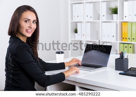 Pretty businesswoman sitting at her workplace, typing at her laptop and looking at the viewer. Concept of office employee portrait