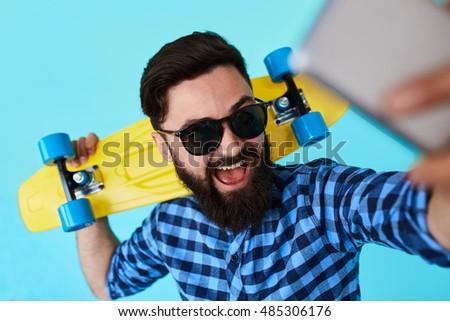 young handsome bearded hipster man makes a selfie on a blue background while holding a skateboard. Stylish male making a self portrait with smartphone