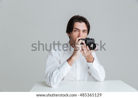 Young bristled man ready to make a photo with camera while sitting at the table isolated on the gray background