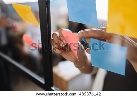 Close up shot of hands of woman sticking adhesive notes on glass wall in office Royalty-Free Stock Photo #485376142