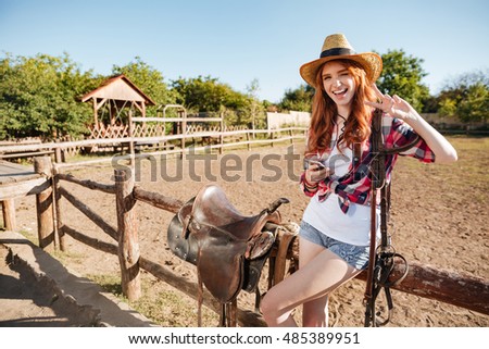 Happy cute young woman cowgirl with cell phone standing and showing peace sign