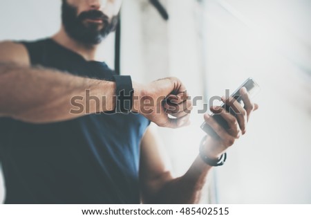 Close-up Shot Bearded Sportive Man After Workout Session Checks Fitness Results Smartphone.Adult Guy Wearing Sport Tracker Wristband Arm.Training hard inside gym.Horizontal bar background.Blurred Royalty-Free Stock Photo #485402515