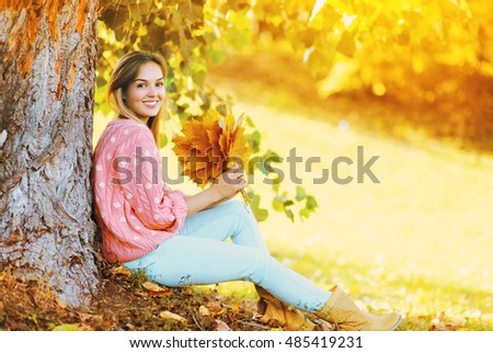 Happy smiling woman with yellow maple leafs sitting under tree in sunny autumn day