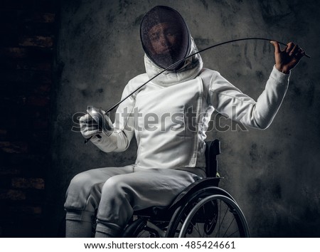 Female fencer in wheelchair with safety mask of a face, holding rapier on grey background.
