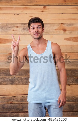 Cheerful young man standing and showing peace sign over wooden background