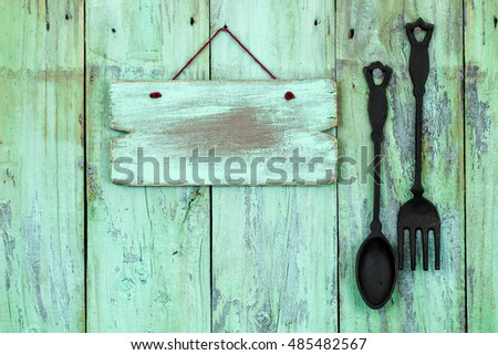 Blank wood sign hanging on antique rustic mint green wood background by black cast iron spoon and fork; restaurant message or menu board