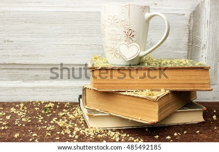Romantic composition with stack of vintage books with very old paper and covers and cup of hot tasty tea in ceramic cup with label in heart shape on wooden background. Copy space