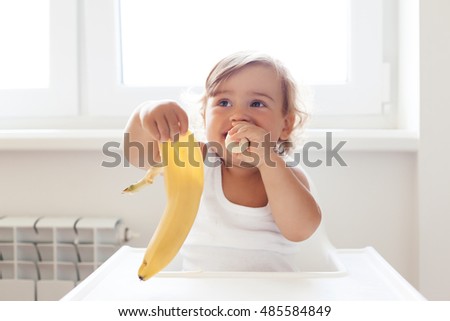 Cute baby 1,4 years old sitting on high children chair and eating fruit alone in white kitchen Royalty-Free Stock Photo #485584849