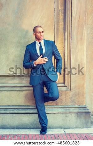 Portrait of Young Strong Businessman. Wearing blue suit, white shirt, black tie, leather shoes, Mixed Race French guy with shaved head stands by vintage wall in New York, thinking. Filtered effect.

