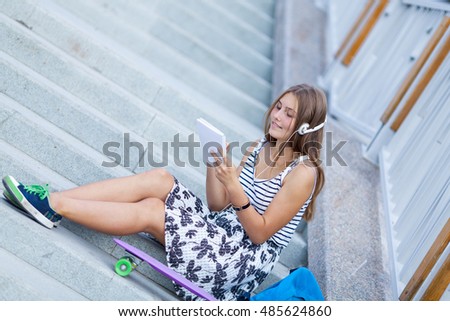 Beautiful, happy young girl with smartphone sitting on stairs. She listens to music on headphones, outdoor, summer