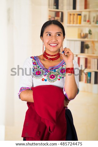 Beautiful young lawyer wearing black skirt, traditional andean blouse with necklace, standing posing for camera, holding red jacket, smiling happily, bookshelves background