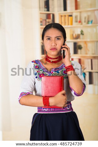 Beautiful young lawyer wearing black skirt, traditional andean blouse with necklace, standing posing for camera, holding red book and talking on phone, serious facial expression, bookshelves