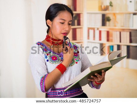 Beautiful young lawyer wearing traditional andean blouse, holding glasses and book reading, bookshelves background