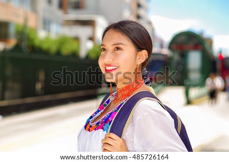 Pretty young woman wearing traditional andean blouse and blue backpack, waiting for bus at outdoors station platform, smiling happily