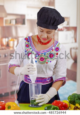 Young woman chef wearing traditional andean blouse and cooking hat, vegetables on desk, using handheld blender, kitchen background