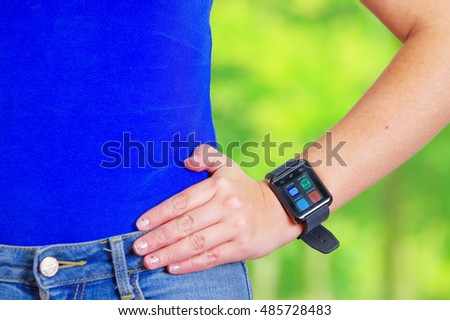 Closeup waist young sporty female wearing blue top with bare arms, smart watch on arm, screen lit up, green forest background