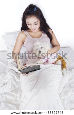 Picture of pretty girl holding book and reading it while resting on bed with her dog, isolated on white background