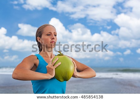 Woman fitness exercise with raw coconut as weight to keep fit and health. Ocean beach surf background. Healthy lifestyle, sport activity and natural flesh on summer family holiday in tropical island.
