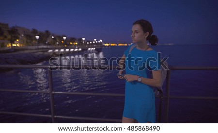 A beautiful woman in the blue dress standing near the sea and looking at night city panorama