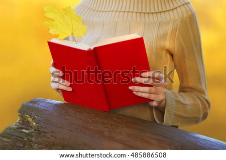 Female hands holding red book with autumn yellow maple leaf closeup over blur background