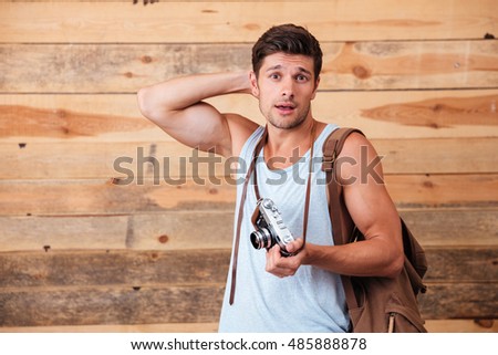 Handsome confused man tourist with backpack and retro camera scratching head isolated on a wooden background