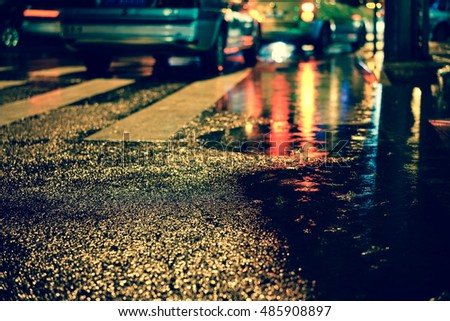 Rainstorm in the big city night, light from the shop windows reflected on the road on which cars travel. View from the level of asphalt