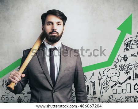 Portrait of businessman in suit holding baseball bat and standing near concrete wall with green arrow and startup images.  Toned image
