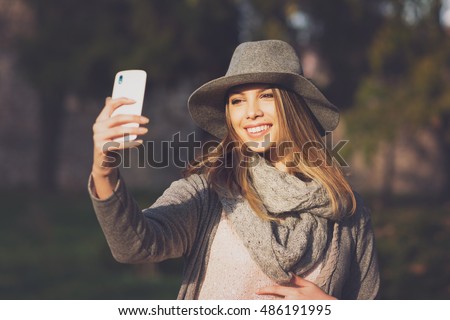 Modern young beautiful woman taking a selfie in park in autumn. Teenage girl in gray fedora hat, gray scarf and sweater photographing herself outdoors in fall. Matte filter applied, natural light.