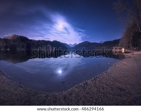 Colorful night landscape with lake, mountains, forest, stars, full moon, purple sky and clouds reflected in water. Spring night in Alpsee lake in Germany. Panoramic photo. Nature background