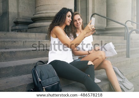 Two female travel bloggers are taking selfie by the camera of a mobile phone while sitting on the stairs of a city square during the weekend journey. Two females are having fun while taking photos